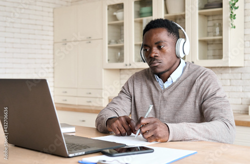 Adult serious African American black male student wearing headphones sitting at kitchen table watching online video conference learning using laptop computer in homeoffice, looking in monitor.