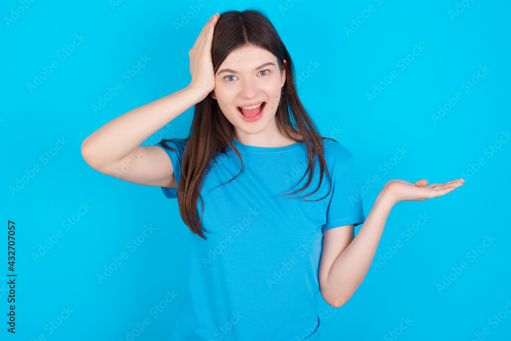 Shocked amazed surprised young beautiful Caucasian woman wearing blue T-shirt over blue wall hold hand offering proposition Shocked amazed surprised young beautiful Caucasian woman wearing blue T-shirt over blue wall hold hand offering proposition
