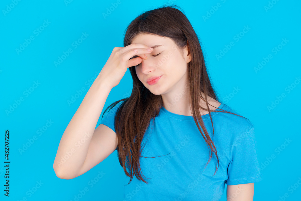 Sad young beautiful Caucasian woman wearing blue T-shirt over blue wall suffering from headache holding hand on her face Sad young beautiful Caucasian woman wearing blue T-shirt over blue wall suffering from headache holding hand on her face