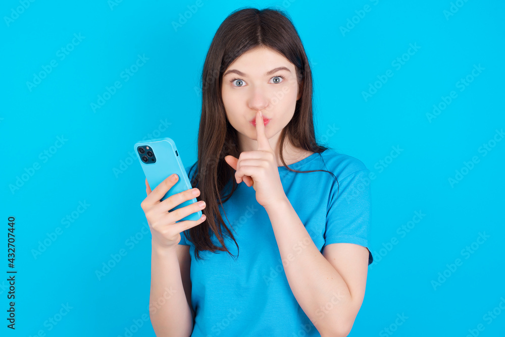 young beautiful Caucasian woman wearing blue T-shirt over blue wall holding modern gadget ask not tell secrets young beautiful Caucasian woman wearing blue T-shirt over blue wall holding modern gadget ask not tell secrets