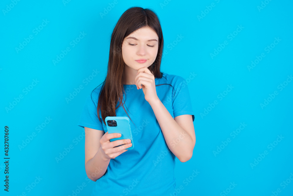 Naklejka premium Thoughtful happy young beautiful Caucasian woman wearing blue T-shirt over blue wall hold look phone, copyspace