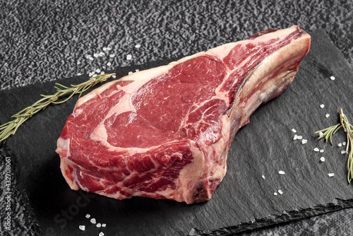 a piece of raw beef steak with a bone lies on a black stone background with a decor of dry greens