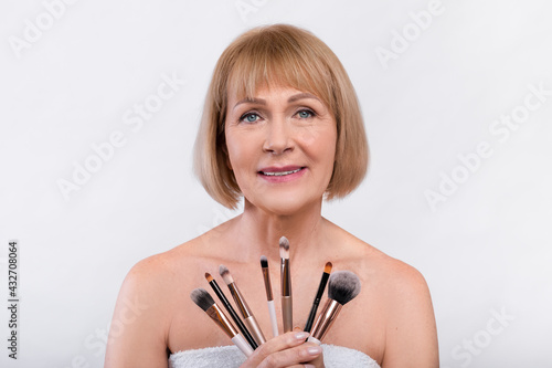Portrait of smiling mature woman holding set of cosmetic brushes over grey studio background