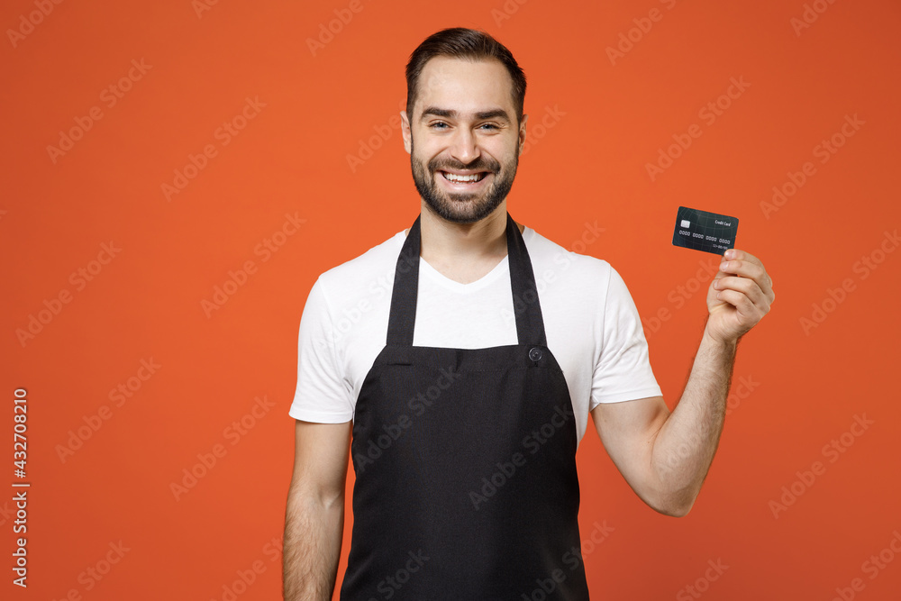 Young successful happy fun man 20s barista bartender barman employee wearing black apron white t-shirt work in coffee shop hold credit bank card isolated on orange background. Small business startup Young successful happy fun man 20s barista bartender barman employee wearing black apron white t-shirt work in coffee shop hold credit bank card isolated on orange background. Small business startup