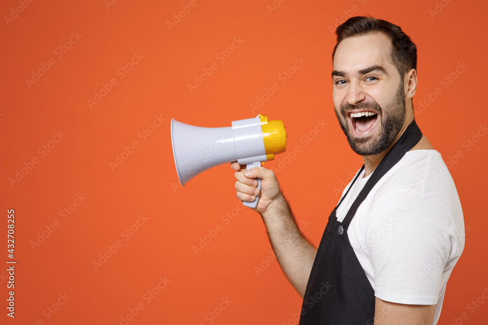 Side view excited young man 20s barista bartender barman employee wearing black apron white t-shirt work in coffee shop scream in megaphone shout isolated on orange background. Small business startup. Side view excited young man 20s barista bartender barman employee wearing black apron white t-shirt work in coffee shop scream in megaphone shout isolated on orange background. Small business startup.