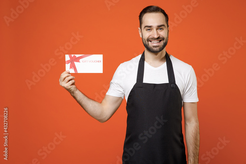 Young happy fun man 20s barista bartender barman employee wearing black apron white t-shirt work in coffee shop hold gift voucher flyer mock up isolated on orange background. Small business startup