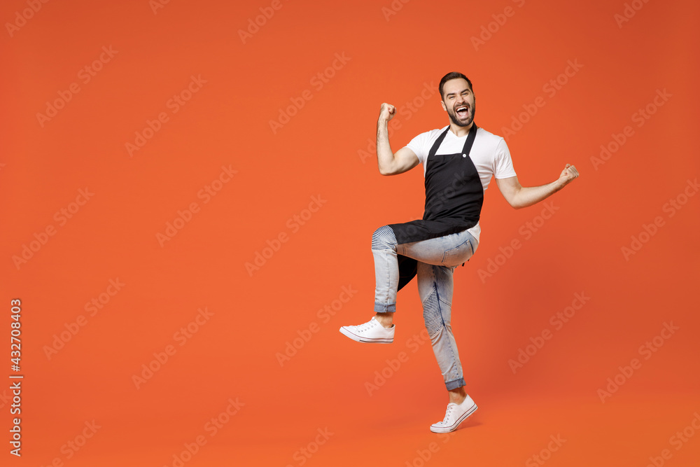 Full length young man barista bartender barman employee in black apron white t-shirt work in coffee shop do winner gesture raised up leg isolated on orange background studio Small business startup
