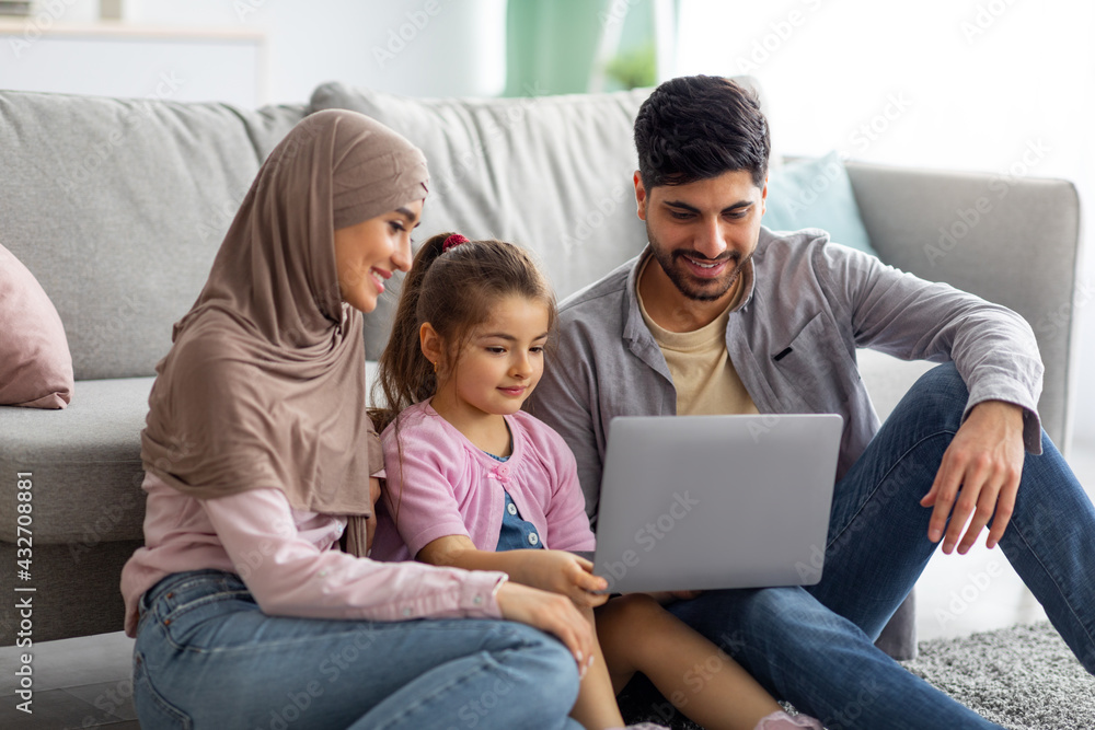 Muslim mother, father and daughter using laptop together, watching family photos while sitting on floor