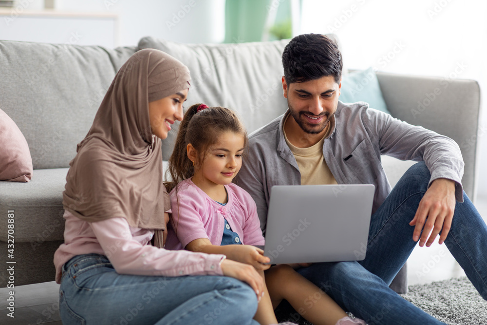 Muslim mother, father and daughter using laptop together, watching family photos while sitting on floor Muslim mother, father and daughter using laptop together, watching family photos while sitting on floor