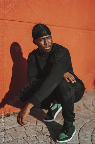 A confident Black man in black casual clothes and do-rag squating against a red wall photo