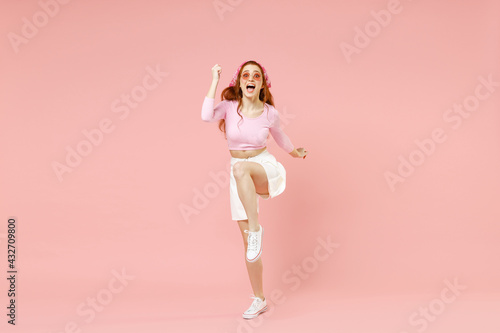 Full length young overjoyed trendy woman in rose clothes bandana glasses do winner gesture clench fist with raised up leg isolated on pastel pink background studio portrait People lifestyle concept.