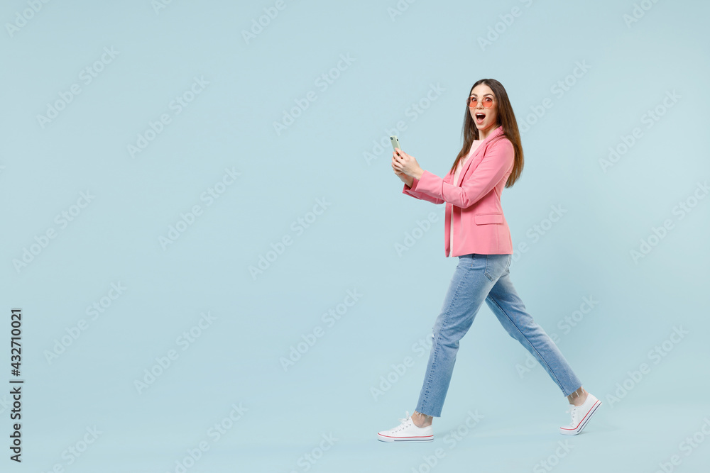 Full length side view young fun smiling happy woman 20s in pastel pink clothes glasses using mobile cell phone chatting online isolated on blue background studio. People lifestyle technology concept Full length side view young fun smiling happy woman 20s in pastel pink clothes glasses using mobile cell phone chatting online isolated on blue background studio. People lifestyle technology concept