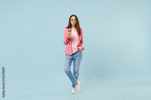 Full length young fun happy smiling satisfied caucasian woman in pastel pink clothes glasses holding in hands paper cup of coffee hot tea isolated on blue background studio. People lifestyle concept.