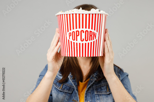 Young caucasian woman 20s in casual denim jacket yellow t-shirt hold hiding covering face with red plastic striped takeaway popcorn bucket isolated on grey background studio People lifestyle concept. photo