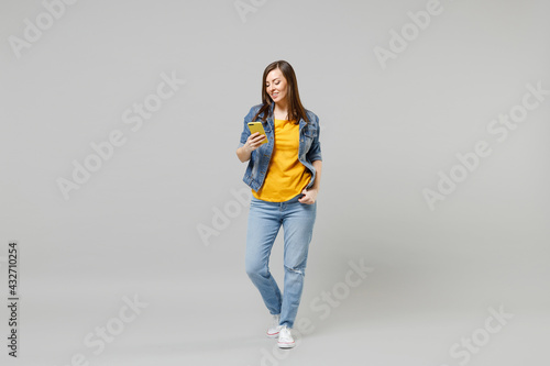 Full length young happy caucasian woman in casual trendy denim jacket yellow t-shirt using mobile cell phone browsing chat online isolated on grey background studio portrait. People lifestyle concept
