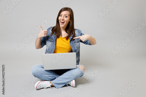 Full length young caucasian woman in denim jacket yellow t-shirt point finger on laptop pc computer browsing sitting cross-legged show thumb up isolated on grey background. People lifestyle concept. © ViDi Studio