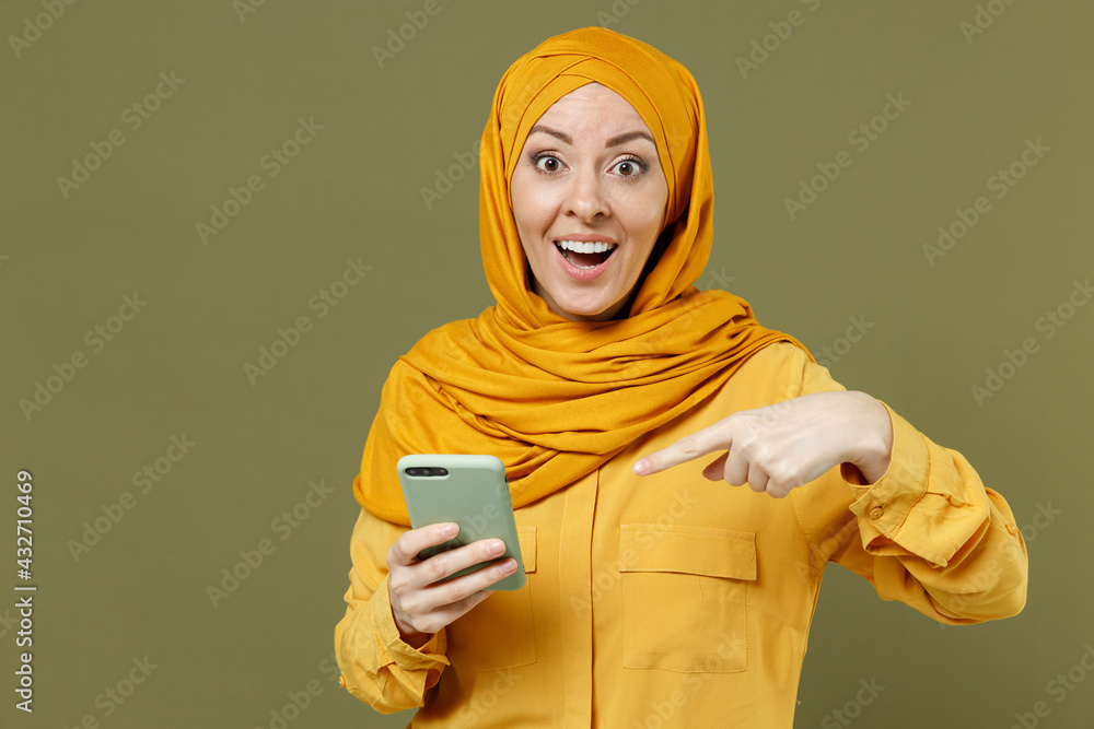 Surprised young arabian asian muslim woman in abaya hijab yellow clothes point index finger on mobile cell phone isolated on olive green background studio portrait. People uae islam religious concept. Surprised young arabian asian muslim woman in abaya hijab yellow clothes point index finger on mobile cell phone isolated on olive green background studio portrait. People uae islam religious concept.