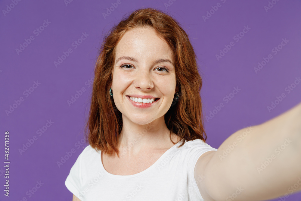 Close up young smiling happy cheerful friendly redhead woman 20s wearing white basic casual t-shirt doing selfie shot on mobile phone looking camera isolated on dark violet color background studio Close up young smiling happy cheerful friendly redhead woman 20s wearing white basic casual t-shirt doing selfie shot on mobile phone looking camera isolated on dark violet color background studio