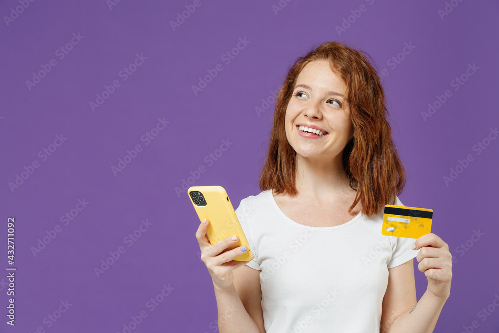 Young smiling happy dreamful woman 20s in white basic casual t-shirt holding using mobile cell phone credit bank card purchasing online look aside isolated on dark violet background studio portrait Young smiling happy dreamful woman 20s in white basic casual t-shirt holding using mobile cell phone credit bank card purchasing online look aside isolated on dark violet background studio portrait