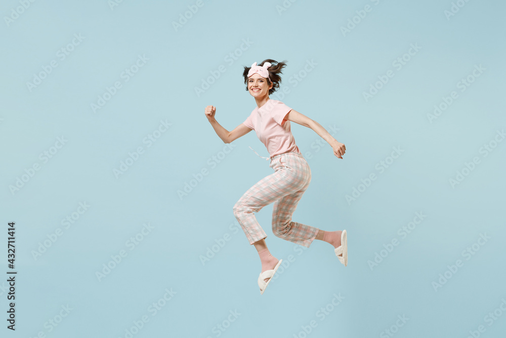 Full length side view sporty young woman in pajamas jam sleep eye mask rest relax at home jump high running fast isolated on pastel blue background studio portrait. Good mood night bedtime concept