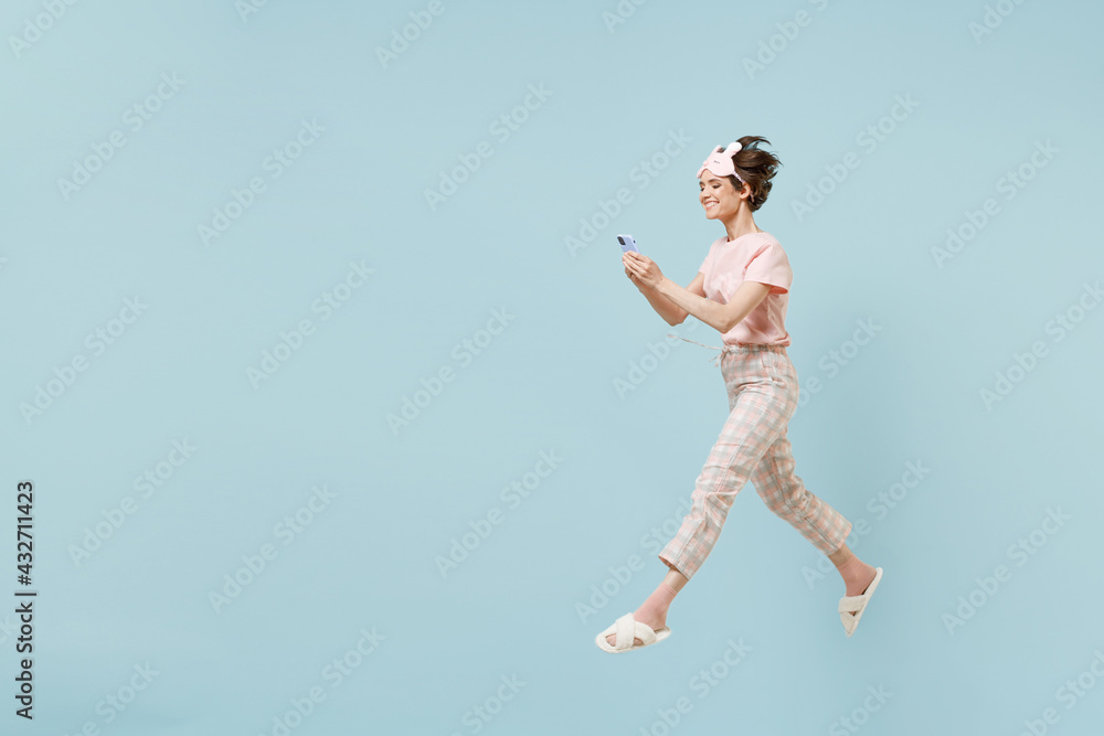 Full length side view young woman 20s in pajamas jam sleep eye mask rest relaxing at home jump high hold mobile cell phone isolated on pastel blue background studio. Good mood night bedtime concept. Full length side view young woman 20s in pajamas jam sleep eye mask rest relaxing at home jump high hold mobile cell phone isolated on pastel blue background studio. Good mood night bedtime concept.
