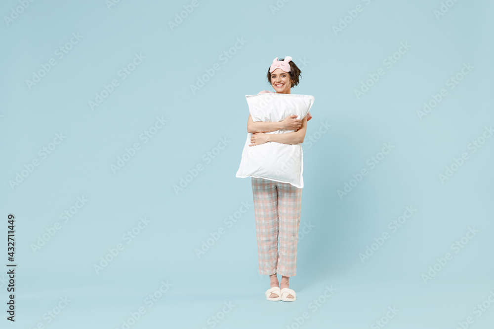 Full length young smiling happy woman 20s in pajamas jam sleep eye mask rest relax at home holding pillow look camera isolated on pastel blue color background studio. Good mood night bedtime concept