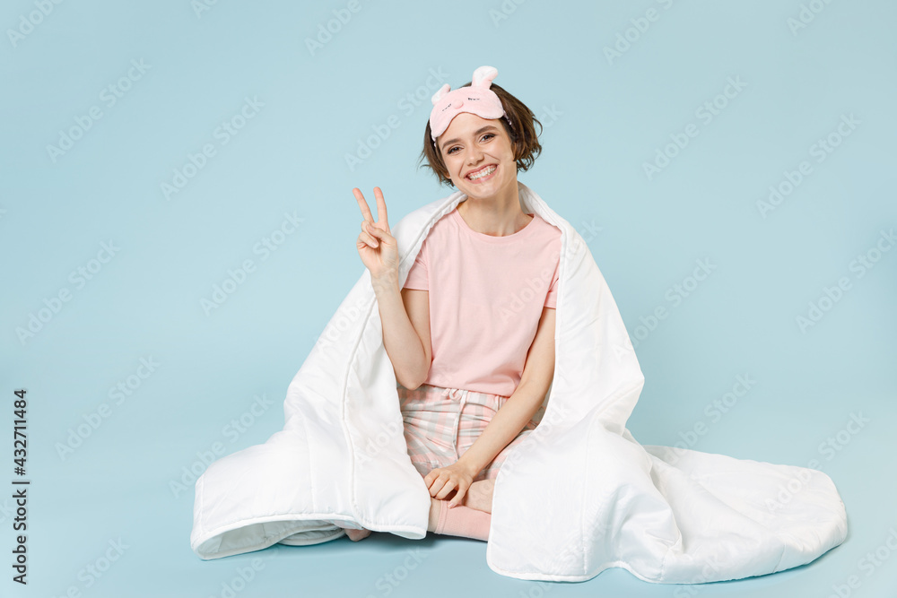 Young fun woman in pajamas jam sleep eye mask rest relax at home sit wrap covered under blanket duvet show victory gesture isolated on pastel blue background studio Good mood night bedtime concept