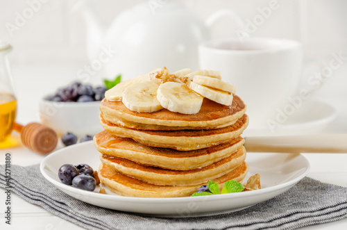 A stack of oatmeal banana pancakes with slices of fresh bananas, walnuts and honey on top with cup of tea on a white wooden background. A healthy breakfast. Copy space.