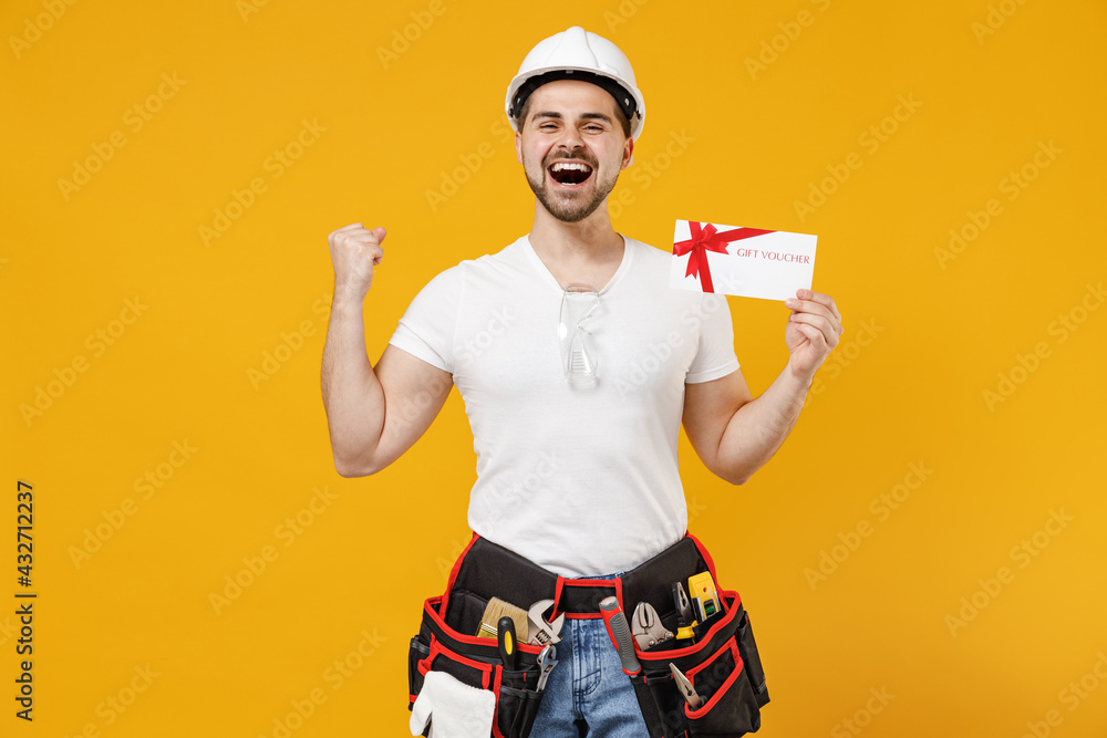 Young smiling employee handyman man in protective helmet hardhat hold gift voucher flyer mock up do winner gesture isolated on yellow background Instruments renovation apartment Repair home concept.
