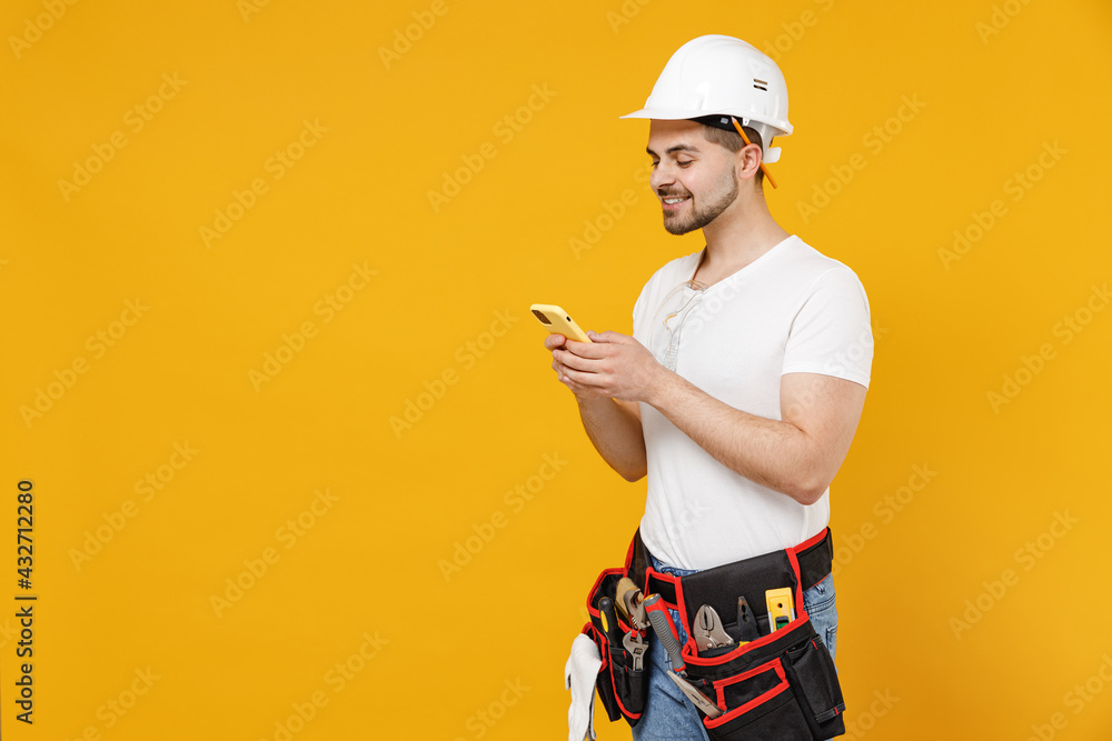 Side view young employee handyman man in protective helmet hardhat use mobile cell phone internet isolated on yellow background. Instruments accessories renovation apartment room. Repair home concept.