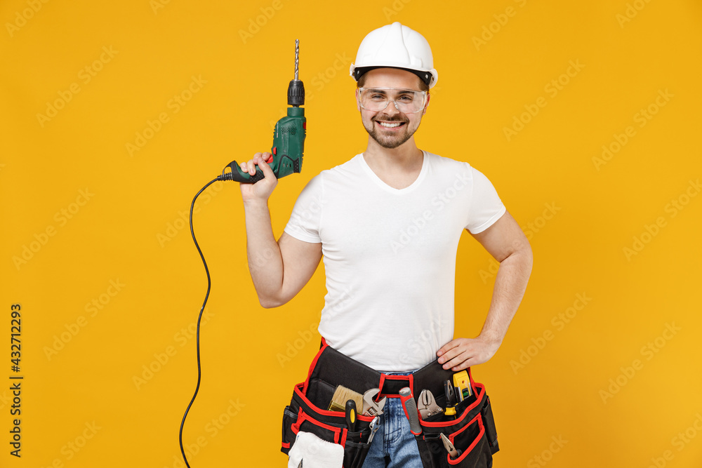 Young fun employee handyman man in protective helmet hardhat glasses holding electric drill isolated on yellow background studio. Instruments accessories renovation apartment room Repair home concept. Young fun employee handyman man in protective helmet hardhat glasses holding electric drill isolated on yellow background studio. Instruments accessories renovation apartment room Repair home concept.