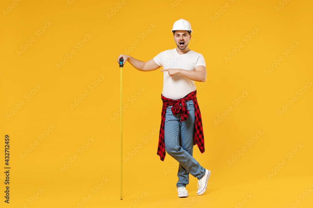 Full length young employee handyman man in protective helmet point finger on measurement tape show thumb up isolated on yellow background Instrument accessory renovation apartment room Repair concept. Full length young employee handyman man in protective helmet point finger on measurement tape show thumb up isolated on yellow background Instrument accessory renovation apartment room Repair concept.