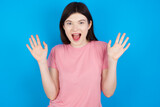 Optimistic young beautiful Caucasian woman wearing pink T-shirt over blue wall raises palms from joy, happy to receive awesome present from someone, shouts loudly, Excited model screaming.