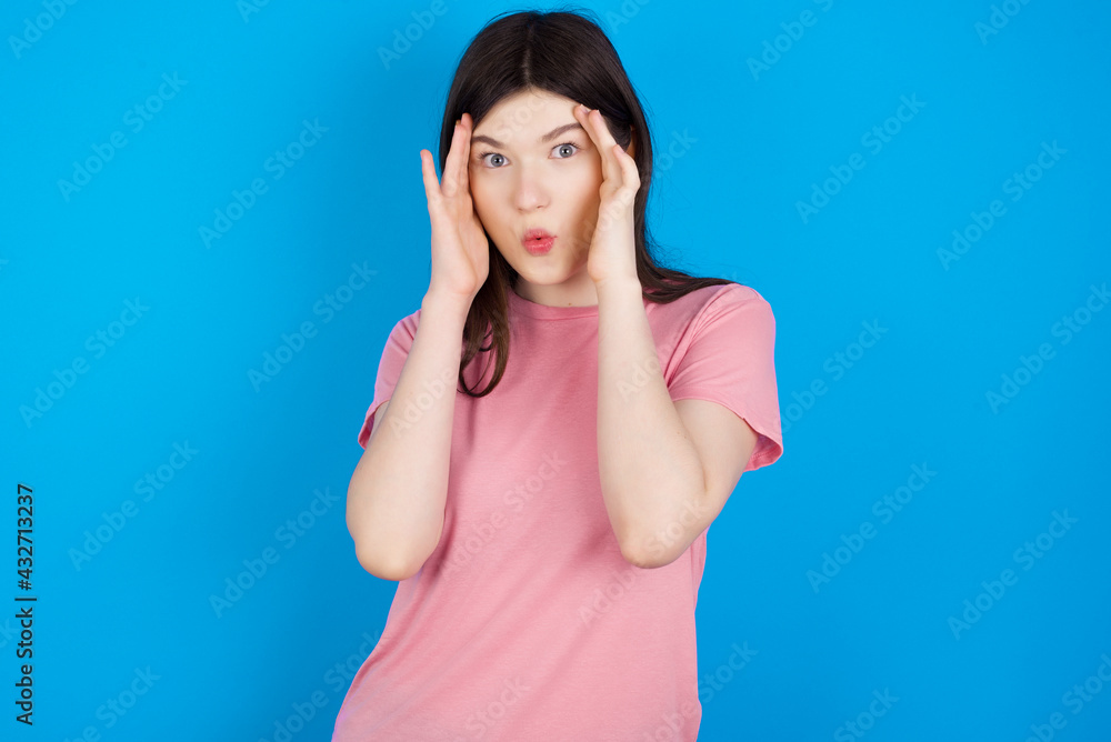 young beautiful Caucasian woman wearing pink T-shirt over blue wall with scared expression, keeps hands on head, jaw dropped, has terrific expression. Omg concept young beautiful Caucasian woman wearing pink T-shirt over blue wall with scared expression, keeps hands on head, jaw dropped, has terrific expression. Omg concept
