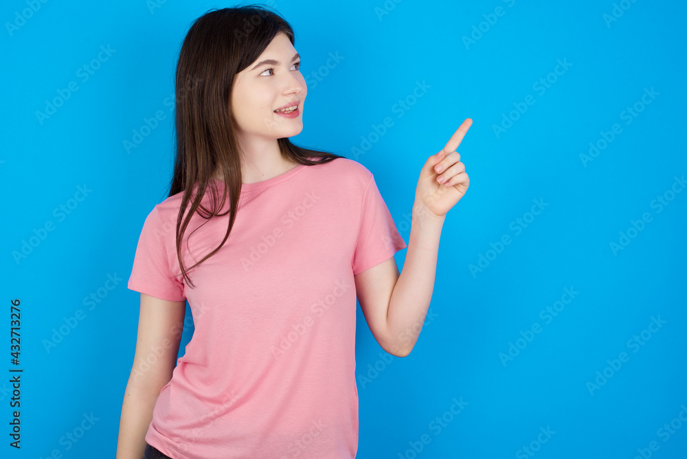 Smiling young beautiful Caucasian woman wearing pink T-shirt over blue wall indicating finger empty space showing best low prices Smiling young beautiful Caucasian woman wearing pink T-shirt over blue wall indicating finger empty space showing best low prices