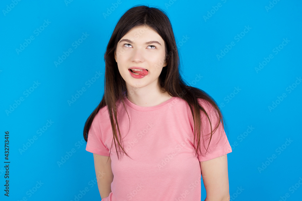 Fototapeta premium young beautiful Caucasian woman wearing pink T-shirt over blue wall showing grimace face crossing eyes and showing tongue. Being funny and crazy
