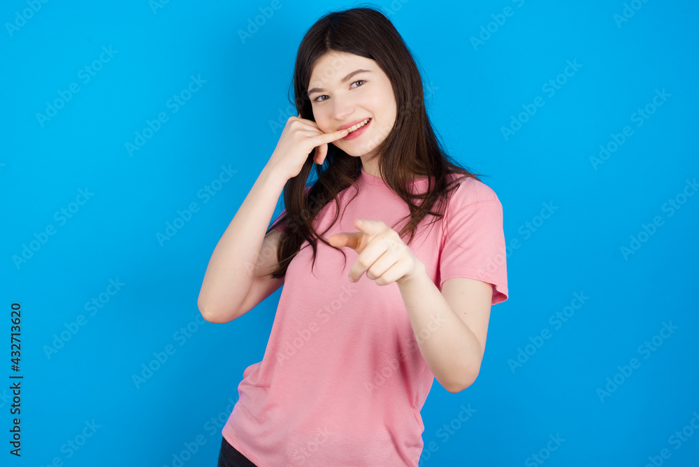 young beautiful Caucasian woman wearing pink T-shirt over blue wall smiling cheerfully and pointing to camera while making a call you later gesture, talking on phone young beautiful Caucasian woman wearing pink T-shirt over blue wall smiling cheerfully and pointing to camera while making a call you later gesture, talking on phone