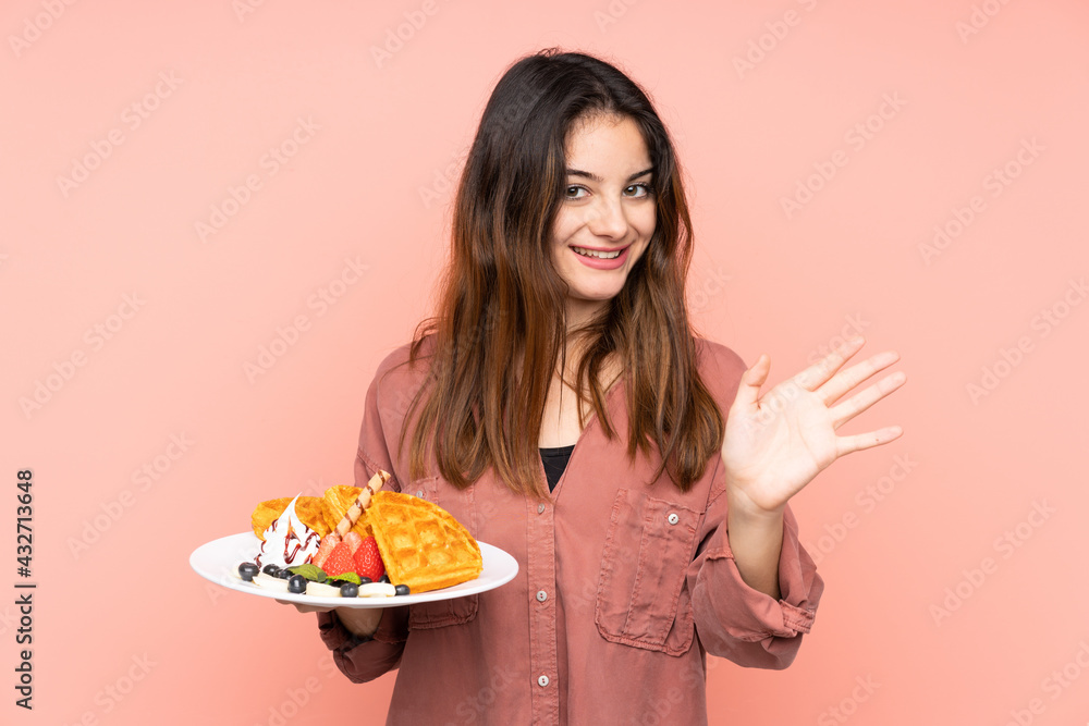Young caucasian woman holding waffles isolated on pink background saluting with hand with happy expression Young caucasian woman holding waffles isolated on pink background saluting with hand with happy expression