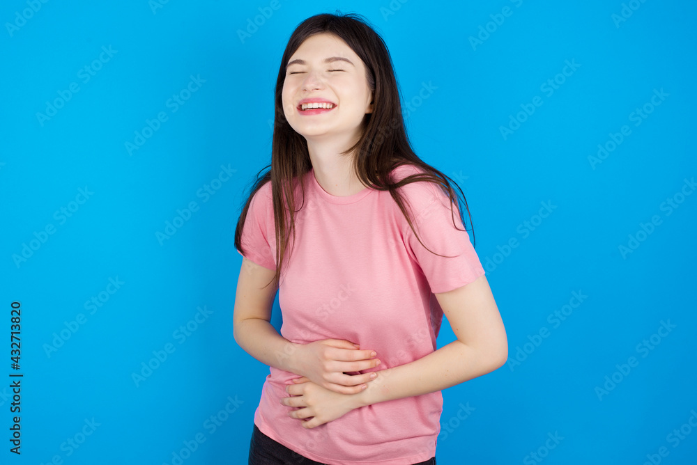 young beautiful Caucasian woman wearing pink T-shirt over blue wall smiling and laughing hard out loud because funny crazy joke with hands on body. young beautiful Caucasian woman wearing pink T-shirt over blue wall smiling and laughing hard out loud because funny crazy joke with hands on body.