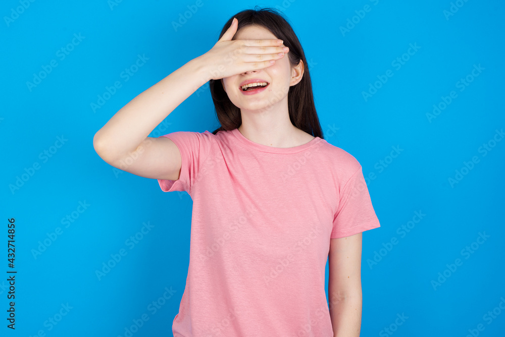 young beautiful Caucasian woman wearing pink T-shirt over blue wall smiling and laughing with hand on face covering eyes for surprise. Blind concept. young beautiful Caucasian woman wearing pink T-shirt over blue wall smiling and laughing with hand on face covering eyes for surprise. Blind concept.