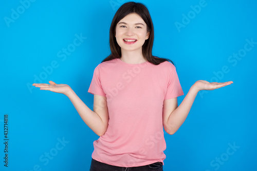 Cheerful cheery optimistic young beautiful Caucasian woman wearing pink T-shirt over blue wall holding two palms copy space
