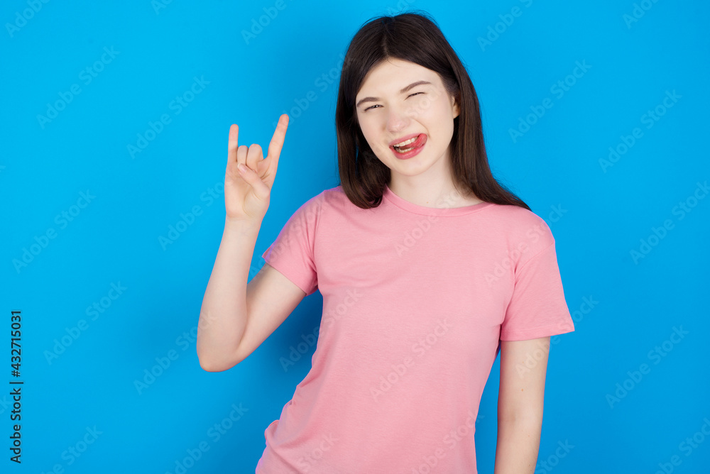 young beautiful Caucasian woman wearing pink T-shirt over blue wall doing a rock gesture and smiling to the camera. Ready to go to her favorite band concert. young beautiful Caucasian woman wearing pink T-shirt over blue wall doing a rock gesture and smiling to the camera. Ready to go to her favorite band concert.