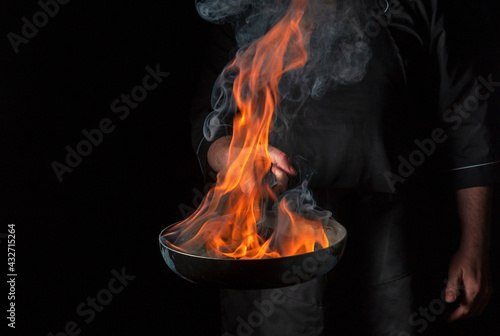  Chef cooking food in pan with fire flame on black background. Restaurant and hotel service concept. Free advertising space