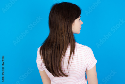 The back side view of a young beautiful Caucasian woman wearing stripped T-shirt over blue wall. Studio Shoot.
