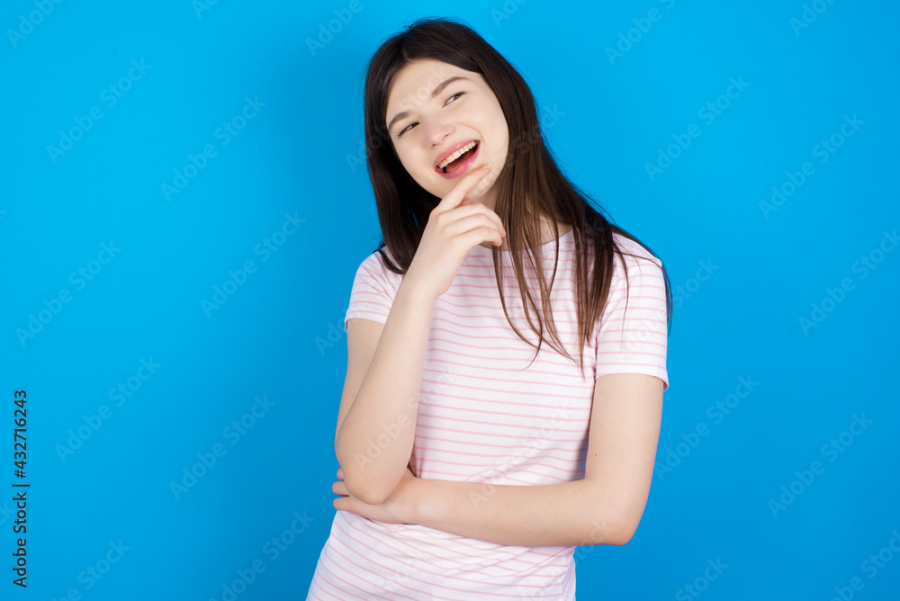 young beautiful Caucasian woman wearing stripped T-shirt over blue wall laughs happily keeps hand on chin expresses positive emotions smiles broadly has carefree expression young beautiful Caucasian woman wearing stripped T-shirt over blue wall laughs happily keeps hand on chin expresses positive emotions smiles broadly has carefree expression