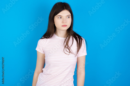 young beautiful Caucasian woman wearing stripped T-shirt over blue wall stares aside with wondered expression has speechless expression. Embarrassed model looks in surprise