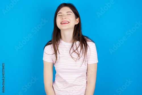Positive young beautiful Caucasian woman wearing stripped T-shirt over blue wall with overjoyed expression closes eyes and laughs shows white perfect teeth