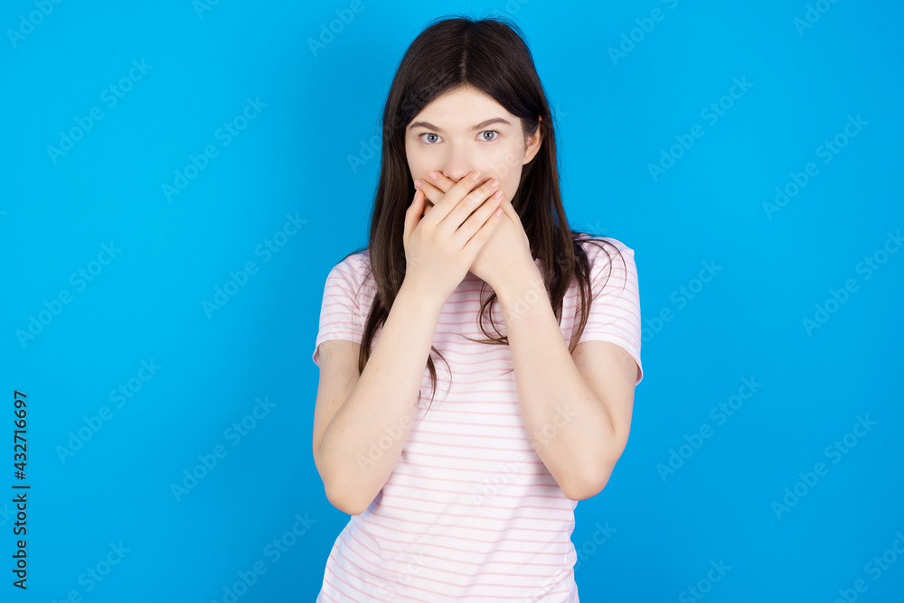Stunned young beautiful Caucasian woman wearing stripped T-shirt over blue wall covers both hands on mouth, afraids of something astonishing Stunned young beautiful Caucasian woman wearing stripped T-shirt over blue wall covers both hands on mouth, afraids of something astonishing