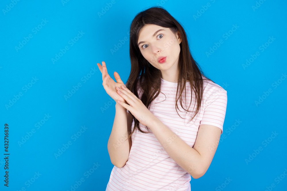 Fototapeta premium Surprised emotional young beautiful Caucasian woman wearing stripped T-shirt over blue wall rubs palms and stares at camera with disbelief