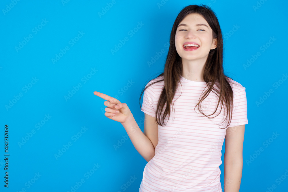 young beautiful Caucasian woman wearing stripped T-shirt over blue wall laughs happily points away on blank space demonstrates shopping discount offer, excited by good news or unexpected sale. young beautiful Caucasian woman wearing stripped T-shirt over blue wall laughs happily points away on blank space demonstrates shopping discount offer, excited by good news or unexpected sale.